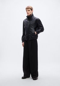 Fekete bőr bombázó dzseki magas gallérral, cipzáras záródással és rugalmas mandzsettákkal, laza fekete nadrággal és fényes cipővel párosítva.