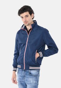 Giacca bomber navy con colletto alto, zip rossa a contrasto e polsini a righe. Materiale liscio e leggero. Due tasche laterali e orlo aderente.