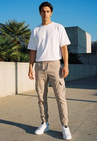 Mladi muškarac stoji na otvorenom noseći bijelu majicu kratkih rukava, bež cargo hlače i bijele tenisice, s palmama i betonskim zidovima u pozadini.