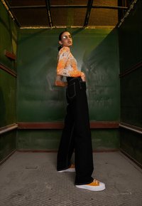 Mustat korkeavyötäröiset farkut valkoisilla ompeleet, yhdistettynä pitkähihaiseen, kuvioituun oranssiin toppiin ja oransseihin lenkkareihin; aseteltu hississä.