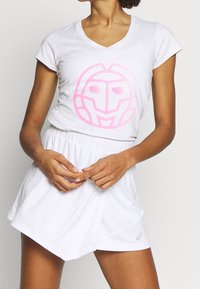 Bílé sportovní tričko s růžovým grafickým motivem ve tvaru masky, doplněné bílou plisovanou sukní. Krátké rukávy a véčkový výstřih.