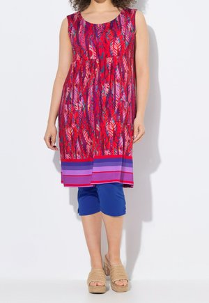 Femme portant une robe sans manches rouge et violette à motifs avec deux poches avant, des leggings capri bleus et des sandales compensées beige.