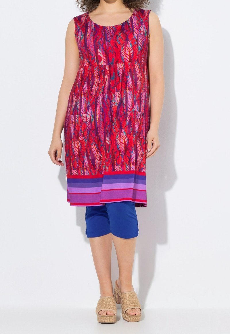 Femme portant une robe sans manches rouge et violette à motifs avec deux poches avant, des leggings capri bleus et des sandales compensées beige.