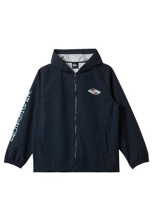 Dreng iført navy lynlås-hættetrøje med logo på bryst og ærme, lyseblå jeans og sorte og hvide sneakers, står mod en hvid baggrund.