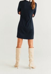 Abito a scacchi navy con silhouette aderente, caratterizzato da un colletto alto e una zip nascosta sul retro, abbinato a stivali crema al ginocchio.