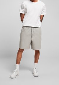 Uomo in piedi con le mani dietro la schiena, indossa una maglietta bianca, pantaloni corti grigi con coulisse, calze bianche e scarpe da ginnastica bianche, su un pavimento grigio.