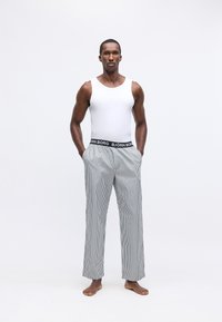 Vīrietis stāv basām kājām, ģērbies baltā bezpiedurkņu kreklā un svītrainās pidžamas biksēs ar 'BJÖRN BORG' jostasvietā uz baltā fona.