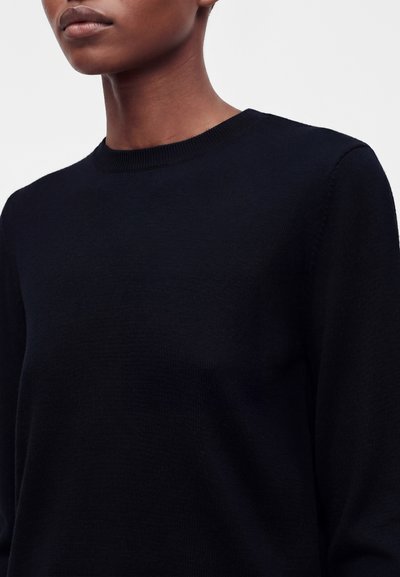 Gros plan d'une personne portant un pull noir uni à manches longues et col rond sur un fond neutre.