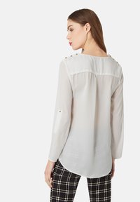Mujer que lleva una blusa blanca transparente de manga larga con botones dorados en los hombros y en las lengüetas de las mangas, combinada con pantalones a cuadros en blanco y negro.