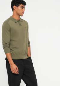 Olijfgroene gebreide polo met een kraag, lange mouwen, ribgebreide manchetten en zoom. Bevat een subtiele logo op de linkerborst. Gedragen met zwarte broek.