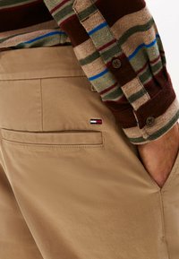 Beige broek met een achterzak, voorzien van een klein rood, wit en blauw logo. De outfit wordt aangevuld met een meerkleurig gestreept overhemd met lange mouwen.