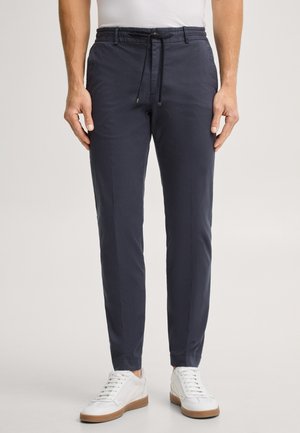 Mann trägt dunkelblaue Slim-Fit-Hose mit Kordelzug in der Taille und weiße Sneakers, steht vor hellem einfarbigem Hintergrund.