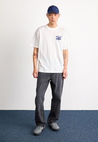 Bela majica s kratkimi rokavi z modrim grafičnim motivom, temno sivi kavbojke in sivi superge. Modra kapica in vidne tetovaže na rokah.