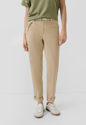 Person trägt beige hochgekrempelte Hose mit geflochtenem Gürtel, olivgrünes Hemd und weiße Sneakers und steht vor einem einfarbigen Hintergrund.