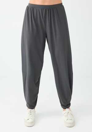Pantaloni larghi grigio scuro con vita elastica indossati con sneakers bianche su una persona in piedi davanti a uno sfondo semplice.