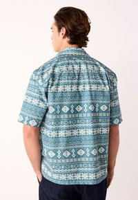 Kortærmet skjorte lavet af tekstureret blåt stof med et hvidt geometrisk mønster og krave, set bagfra.