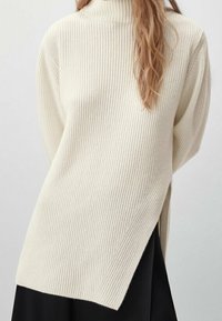 Femme portant un pull côtelé crème à col roulé avec une fente latérale sur une jupe noire, se tenant devant un fond clair.