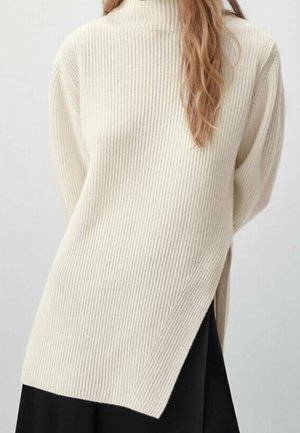 Woman wearing a cream ribbed turtleneck sweater with a side slit over a black skirt, standing against a plain light background.