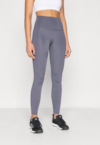 Leggings taille haute gris en matière extensible avec une texture lisse et des coutures ton sur ton. Portés avec des chaussures de sport noires.