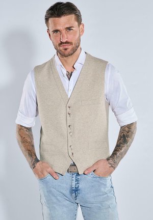 Ein tätowierter Mann trägt eine beigefarbene Knopfveste über einem weißen hochgekrempelten Hemd und hellblauen Jeans, die Hände in den Taschen, vor einem schlichten Hintergrund.