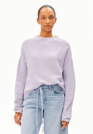Femme portant un pull violet clair et un jean taille haute bleu clair avec un cordon noué à la taille, debout sur fond blanc.