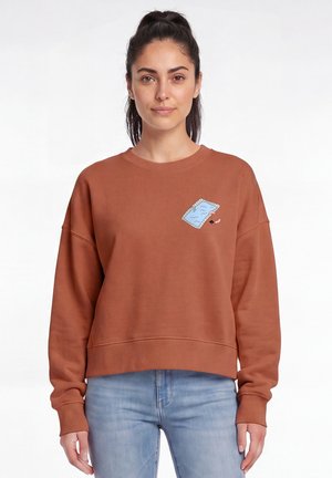 Vrouw met donker haar in een paardenstaart draagt een bruin sweatshirt met een kleine blauwe zwembadgrafiek en lichtblauwe spijkerbroek, staand tegen een witte achtergrond.