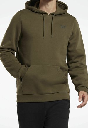 Mann trägt olivgrünen Reebok-Hoodie mit Fronttasche und schwarze Hose, Hand teilweise in der Tasche vor weißem Hintergrund.