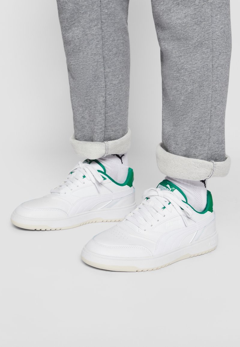 Baskets blanches avec des accents verts, dotées d'une tige en cuir lisse, d'un bout rond, d'un logo embossé et d'une semelle en caoutchouc texturée. Pantalons de survêtement gris avec des ourlets côtelés.