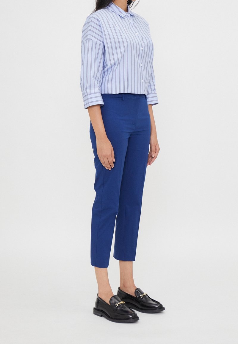 Personne portant un pantalon bleu court, une chemise blanche avec des rayures verticales bleues et des mocassins noirs, debout devant un fond uni.