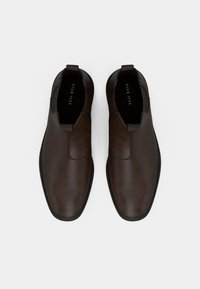 Un paio di scarpe slip-on in pelle marrone con suole nere, pannelli laterali elasticizzati e linguette, viste dall'alto su uno sfondo bianco.