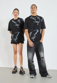 Sorte oversized t-shirts har store grå sommerfugletryk og teksten "TAKE FLIGHT." Den mandlige model bærer slidte sorte jeans og sneakers.