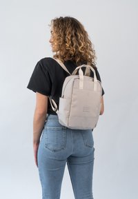 Beige hátizsák vállpántokkal és felső fogantyúkkal. Simár, matt felület, elülső cipzáros zsebbel és logó részlettel. Ívelt forma, kompakt kialakítás.