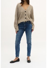 Cardigan lavorato a maglia beige con scollatura a V profonda e cinque bottoni scuri, abbinato a jeans skinny blu medio. Ballerine nere con dettagli a fiocco.