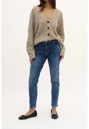 Beige gestrickte Strickjacke mit tiefem V-Ausschnitt und fünf dunklen Knöpfen, kombiniert mit mittelblauen Skinny-Jeans. Schwarze Ballerinas mit Schleifenakzenten.