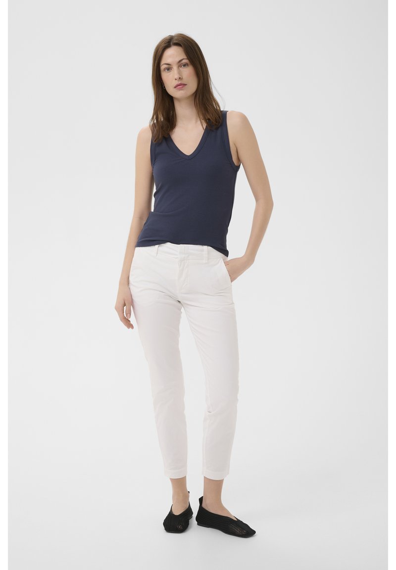 Femeie îmbrăcată într-un top bleumarin fără mâneci, cu decolteu în V, pantaloni albi slim și pantofi slip-on negri, stând cu o mână în buzunar.