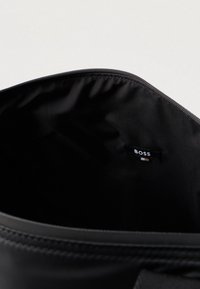 Otwarta czarna torba ukazująca gładkie wewnętrzne wykończenie z małą przyszytą metką z napisem "BOSS" blisko górnej krawędzi oraz widocznym zamkiem błyskawicznym.