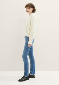 Kreemist ribakudumist kõrge kaelusega sviiter, mida on paaritatud helesiniste sirgete pükstega ja mustade nöörketsidega, seistes vasakule poole neutraalse tausta ees.