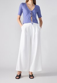Fialový pletený top s výstřihem do V a detaily na zavazování, spárovaný s širokými bílými kalhotami a černými sandály na podpatku. Texturovaná tkanina a upravený střih.