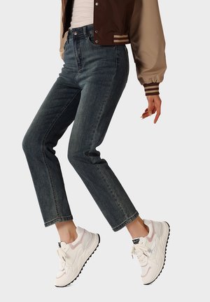 Persona che indossa jeans slim blu scuro, sneakers bianche e rosa, giacca varsity beige e marrone, in posa a metà passo contro uno sfondo neutro.