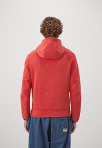 Röd huvtröja med textilier, raglanärmar och ribbad nederkant. Inkluderar en känguruficka och en avslappnad passform.