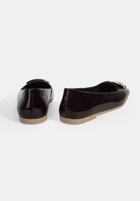 Ballerines noires en cuir verni avec un bout rond, une petite semelle en caoutchouc beige et un accent décoratif doré à l'avant.