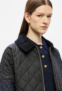 Μαύρο μπουφάν με ραμμένη επένδυση, γιακά από βελούδο σε ναυτικό μπλε, κουμπιά χρυσού χρώματος σε σχήμα τριαντάφυλλου και υφασμένο πουλόβερ σε ναυτικό μπλε από κάτω.