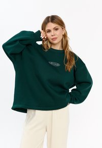 Felpa verde oversize con polsini a coste e scollatura rotonda, caratterizzata da un logo in rilievo bianco sul petto.