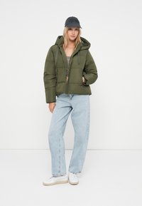 Tânără îmbrăcată cu o geacă puffer verde măsliniu, blugi largi de culoare albastru deschis, șapcă neagră și adidași albi, stând în fața unui fundal alb simplu.