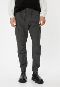 Grijze cargobroek met een elastische tailleband, verstelbaar trekkoord, zijzakken en opgerolde enkels, gecombineerd met zwarte laarzen.