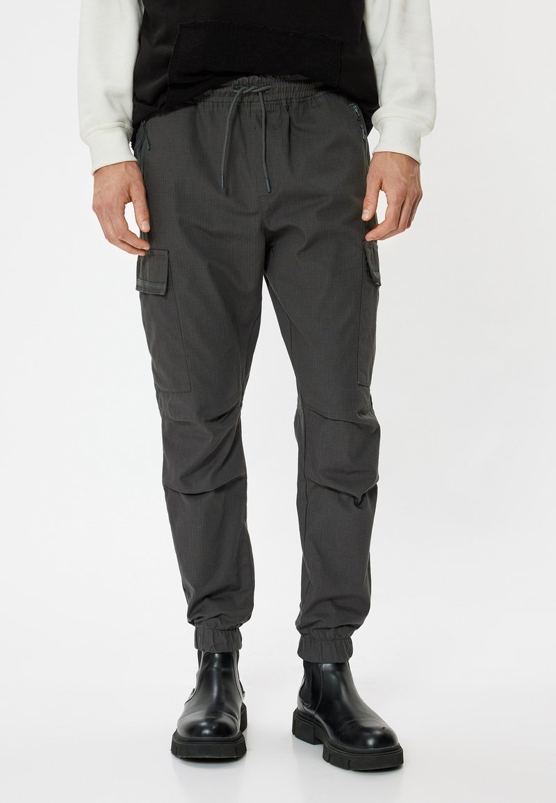 Grijze cargobroek met een elastische tailleband, verstelbaar trekkoord, zijzakken en opgerolde enkels, gecombineerd met zwarte laarzen.