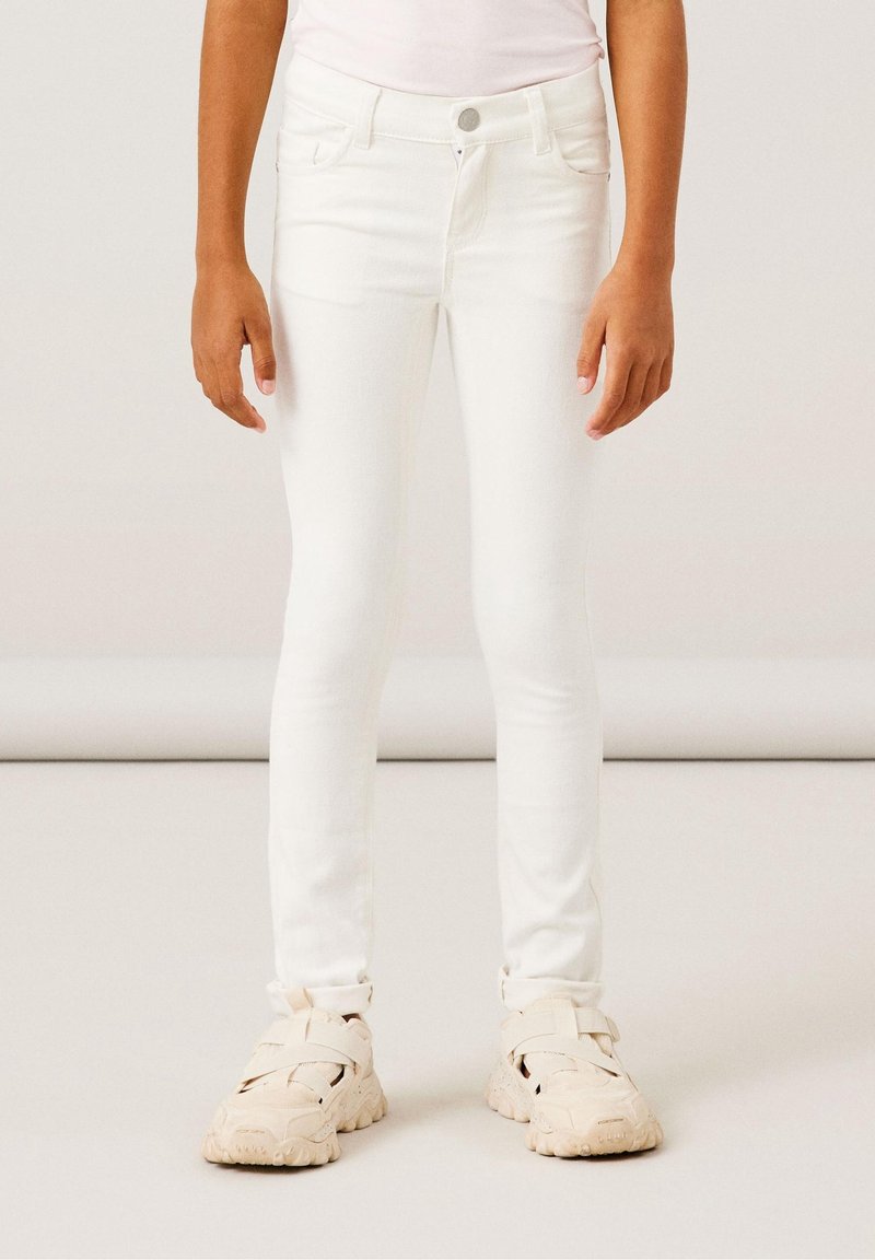 Personne portant un pantalon blanc slim et des baskets beiges épaisses, debout avec les bras détendus le long du corps sur un fond clair.