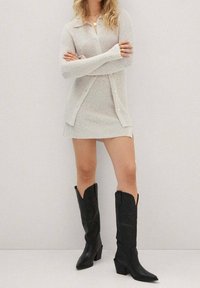 Donna in piedi con le braccia incrociate, indossa un vestito e un cardigan in maglia beige chiaro, abbinati a stivali cowboy neri con tacco e altezza fino al polpaccio, davanti a una parete semplice.
