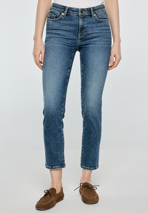 Kobieta ubrana w niebieskie obcisłe jeansy z podwyższonym stanem oraz brązowe mokasyny zamszowe na sznurowadła, stojąca na jednolitym jasnym tle.