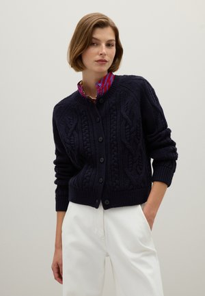 Marine gebreide cardigan met kabelpatroon, ronde halslijn, knoopsluiting aan de voorkant en geribbelde manchetten. Gecombineerd met witte broek.
